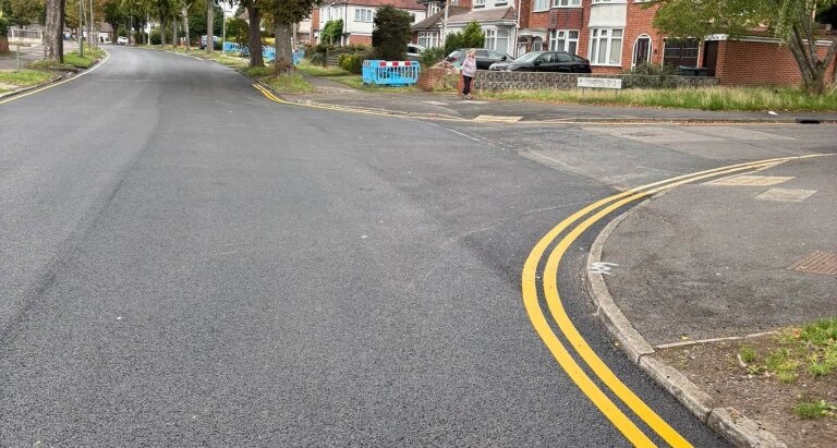 New double yellow lines painted along a road bend in Sheldon on freshly laid dark asphalt, with residential houses visible in the background